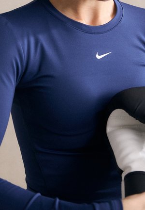 Osoba w granatowej koszulce z długim rękawem Nike trzyma czarno-białą rękawicę bokserską blisko klatki piersiowej.