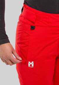 Pantalons de ski rouges avec une texture lisse, dotés d'un ajustement à la taille avec Velcro, d'une poche zippée et d'un logo Millet blanc sur le devant.