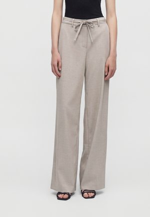 Beige wijde broek met ceintuur in de taille, gedragen met een zwart mouwloos topje en zwarte open-toesandalen tegen een effen achtergrond.