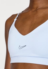 Ανοιχτό μπλε αθλητικό σουτιέν με σχέδιο σε σχήμα V στο ντεκολτέ, κατασκευασμένο από λείο υλικό. Διαθέτ ει ασημένιο λογότυπο Nike μπροστά και ρυθμιζόμενους ιμάντες.