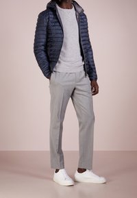 Marinblå vadderad jacka, grå tröja, ljusgrå byxor och vita sneakers. Har quiltad textur och hel dragkedja, med huva.