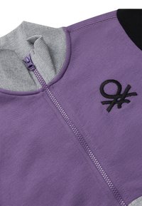 Sudadera morada con cremallera y cuello acanalado gris, con un logo negro bordado. Textura suave con un detalle negro en el hombro que contrasta.