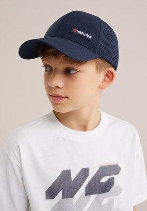 Jeune garçon regardant sur le côté, portant une casquette perforée bleu marine étiquetée "Neolite 5" et un t-shirt blanc avec un texte audacieux "NG".