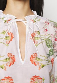 Blouse blanche à imprimé floral dans des tons de rouge et de vert. Dotée d'un col en V profond et d'un cordon à nouer au niveau du col. Tissu léger et aérien.