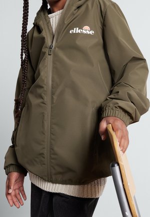 Personne portant une veste ellesse vert olive par-dessus un pull crème, tenant un skateboard en bois à ses côtés sur un fond gris clair.