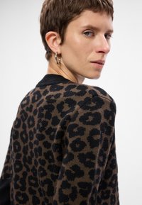 Brązowy sweter w panterkę z czarnymi akcentami, o teksturowanej tkaninie, okrągłym dekolcie i prążkowanych mankietach, prezentujący swobodny i nowoczesny design.