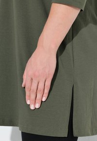 Kortärmad olivgrön tunika, gjord av mjukt tyg, med sidofickor, en slät struktur och sömnadsdetaljer.