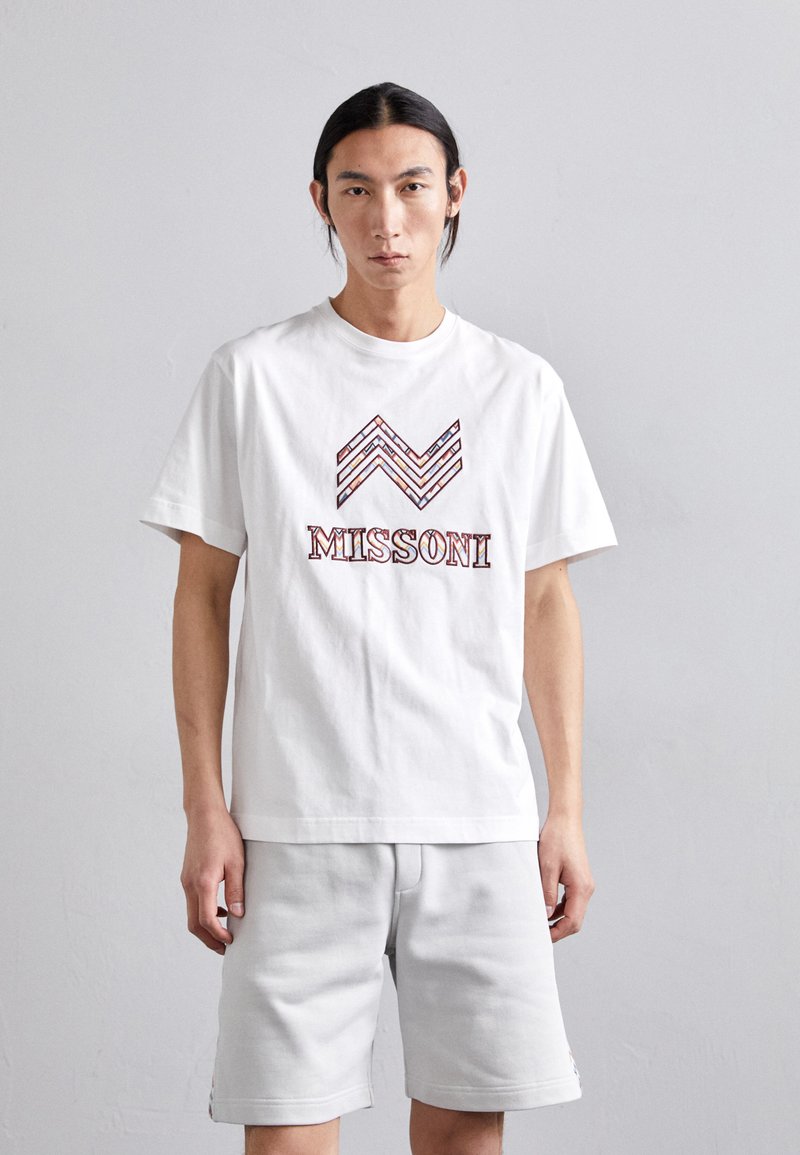 T-shirt en coton blanc avec un design en zigzag dans des tons rouges et marron, avec le logo "MISSONI" affiché en évidence en dessous. Associé à un short gris.