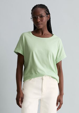 Femme aux cheveux tressés portant de grandes lunettes, un t-shirt vert clair à manches courtes et un pantalon blanc, debout devant un fond uni.