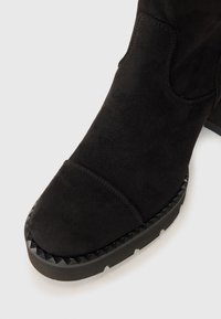 Black suede ankle boot with a rounded toe, featuring a textured studded black rubber sole and visible stitching along the upper.