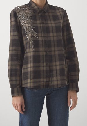 Brązowa i czarna koszula w kratę na guziki z długimi rękawami i srebrnym haftem piór koralikami na lewym ramieniu, noszona z niebieskimi dżinsami.