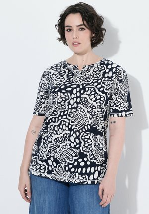 Frau mit kurzen lockigen Haaren, die ein schwarz-weiß gemustertes kurzärmeliges Oberteil und blaue Jeans trägt, steht vor einem weißen Hintergrund.