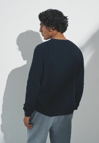 Mornarica pletena majica s opuštenim krojem i rebrastim manžetama i rubom, uparena sa sivim trenirkama. Sjena vidljiva na zidu.