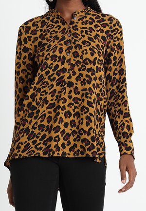 Kvinde iført en langærmet, sennepsgul bluse med sort og mørkerødt leopardmønster og sorte bukser.