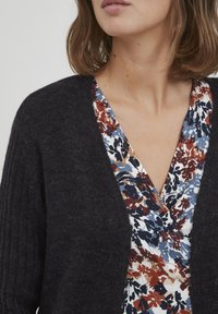 Mörkgrå stickad cardigan med ribbad textur, bärs över en blus med blommönster i blå, röd och vit nyanser och med V-ringning.