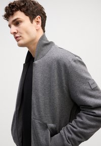 Giacca bomber grigia in morbido tessuto, con collo e polsini a coste, tasche laterali e una patch con logo sulla manica sinistra.