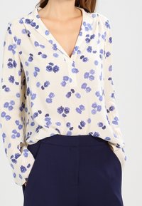 Blouse en crème clair avec un motif floral violet, décolleté en V et manches longues, associée à un pantalon bleu marine. Texture de tissu lisse.