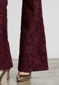 Pantalones acampanados en color granate, confeccionados con tela texturizada y detalle de estampado floral. Presentan un tacón dorado liso y brillante. Fondo de superficie plana.