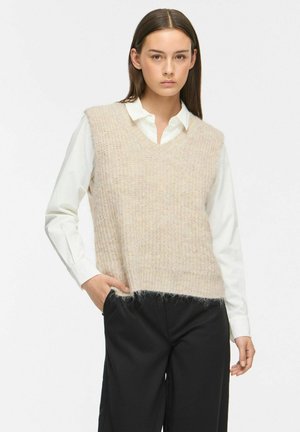 Jeune femme portant un gilet tricoté beige sur une chemise blanche à col et un pantalon noir, debout avec une main dans la poche.