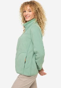 Veste en polaire verte avec un col montant, poches zippées et une texture douce, présentant des détails de couture et une coupe ergonomique.
