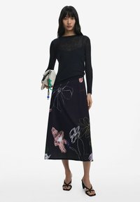 Black long-sleeve top with sheer texture, paired with a black maxi skirt featuring colorful floral patterns. Model holding a light-colored clutch.