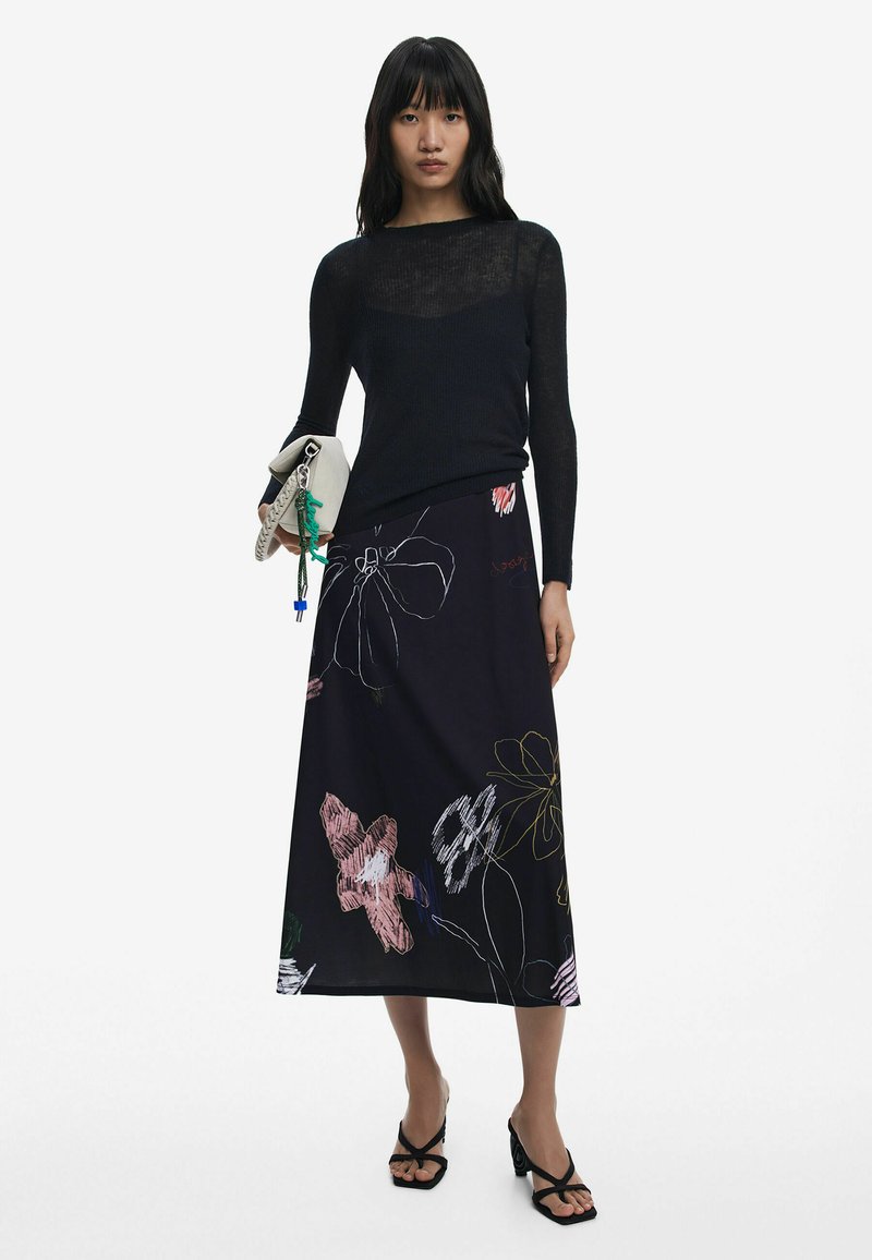 Black long-sleeve top with sheer texture, paired with a black maxi skirt featuring colorful floral patterns. Model holding a light-colored clutch.