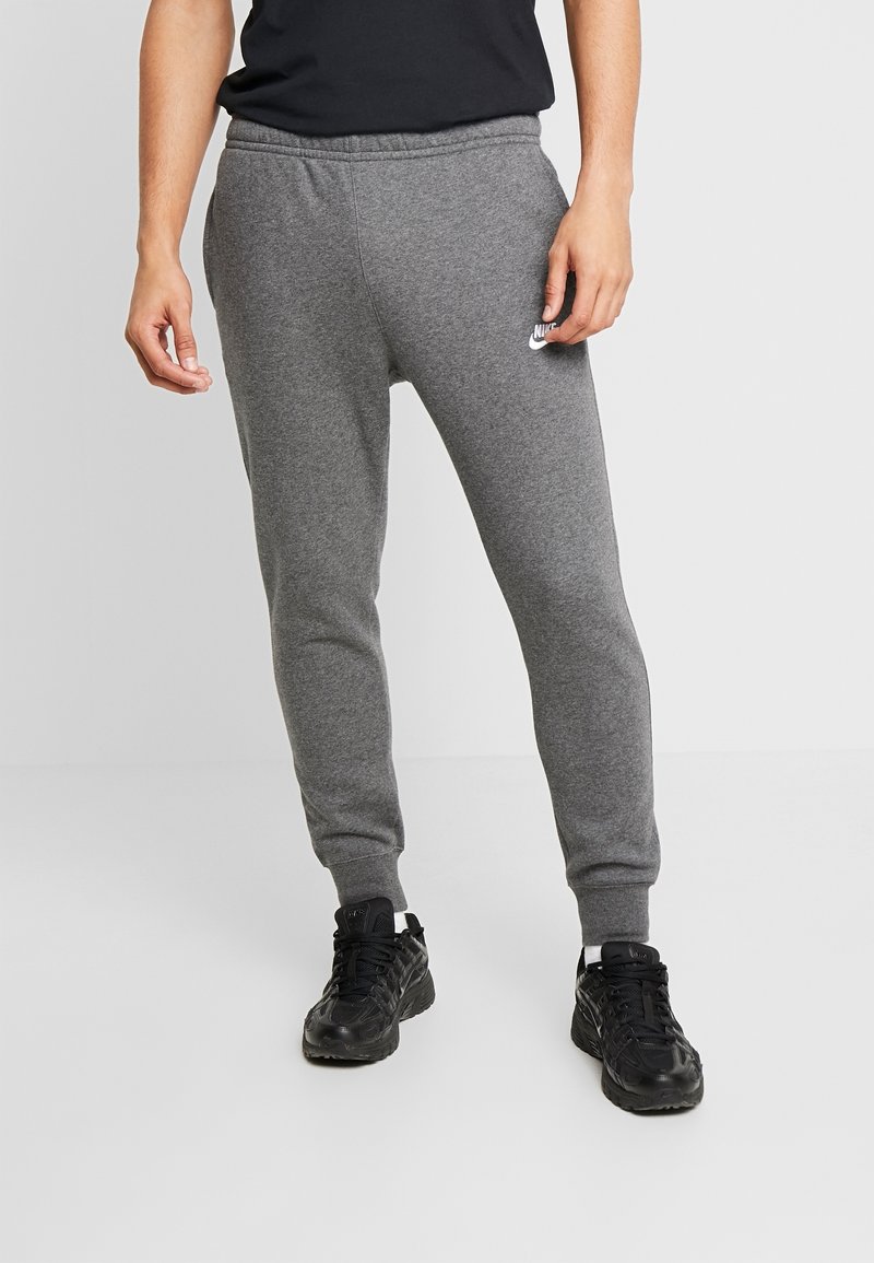 Man som bär grå Nike sweatpants och svarta sneakers står mot en enkel ljus bakgrund.