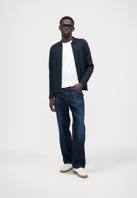 Conjunto casual com um cardigã azul-marinho, t-shirt branca, jeans escuros e sapatilhas brancas com solas castanhas, em fundo simples.