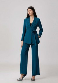 Femme aux longs cheveux bruns portant un tailleur ajusté teal avec ceinture et des escarpins multicolores, se tenant devant un fond uni.