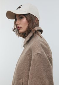 Veste en mélange de laine marron avec un col, comportant une poche latérale. Casquette beige clair avec des accents de logo noirs. Vue de profil du modèle sur fond blanc.