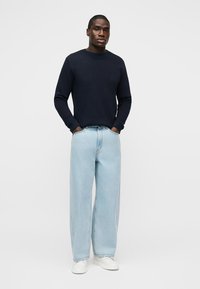 Άνδρας που φοράει μαύρο πουκάμισο με μακριά μανίκια, ανοιχτόχρωμα τζιν με φαρδιά πόδια, λευκά αθλητικά παπούτσια, στέκεται με τα χέρια στις τσέπες μπροστά σε ένα απλό φόντο.