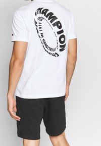 Hvid bomulds T-shirt med stort sort cirkulært logo på ryggen, der står "Champion Established in 1919." Sammen med sorte shorts.