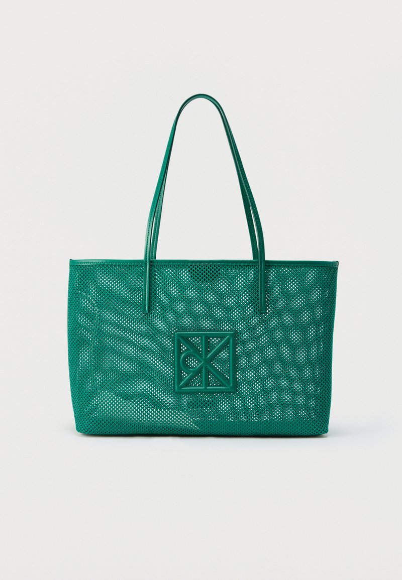Sac fourre-tout en cuir vert avec un design perforé et un patch logo géométrique, comprenant deux bretelles, sur un fond uni.