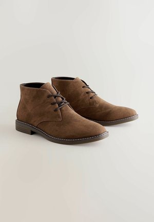 Braune Wildleder-Ankle Boots mit abgerundetem Zehenbereich, Schnürdesign und kontrastierenden weißen Nähten an der Sohle. Flacher Absatz und glattes Innere.