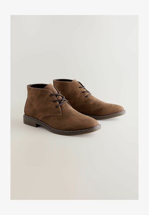 Rjave nubuk škornje do gležnjev z zaobljenim nosom, vezalnim dizajnom in kontrastnim belim šivanjem na podplatu. Ravna peta in gladek notranji del.