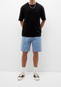 Černé tričko s krátkým rukávem a kapsou, světle modré džíny šortky, bílé ponožky a černé tenisky s krémovými akcenty. Jednoduché pozadí.