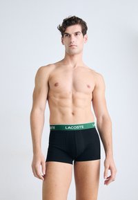 Een naakte man met kort krullend haar die zwarte Lacoste boxershorts met een groene tailleband draagt, terwijl hij tegen een effen lichte achtergrond staat.