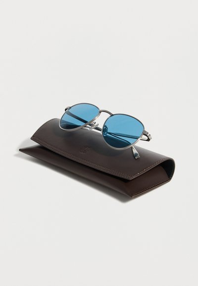 Στρογγυλά γυαλιά ηλίου με ασημένιο πλαίσιο και μπλε φακούς που ακουμπούν σε κλειστή σκούρα καφέ δερμάτινη θήκη σε λευκό φόντο.