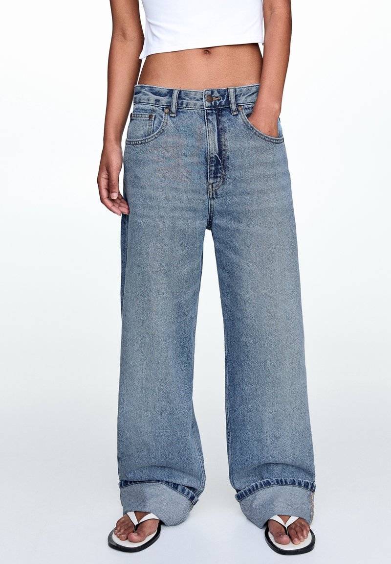 Jeans ampi di colore blu chiaro con vita alta, tasche frontali e orli arrotolati, abbinati a infradito bianche e nere.