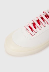 Hvit lær sneakers med rund tå, gjennomsiktig gummisåle og røde tekstilskolisser over en teksturert tunge. Synlig dekorativ søm.