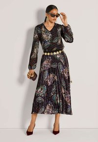 Langdrog met lange mouwen en een V-hals, met een paisley print in zwart, paars en groen. Een gedetailleerde riem en bordeauxrode hakken maken de look compleet.