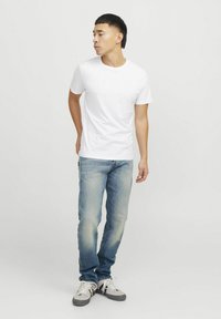 Camiseta blanca de manga corta combinada con jeans de color azul claro. El modelo lleva zapatillas grises y blancas, de pie frente a un fondo liso.