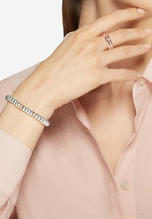 Hand mit einem roségoldenen Doppelbandring nahe am Gesicht, Handgelenk mit einem Armband aus abwechselnden Silber- und Roségoldperlen, weiches rosa Hemd mit Knöpfen.