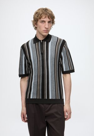 Ung mand med kort krøllet hår iført en sort, grå, lyseblå og beige stribet kortærmet strikket polo shirt og mørke bukser.