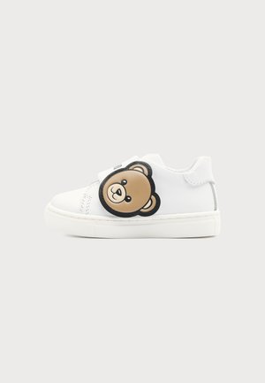 Weiße Kindersneakers mit einem braunen Teddybär-Gesichtsflicken an der Seite und einer dicken weißen Sohle vor einfachem Hintergrund.