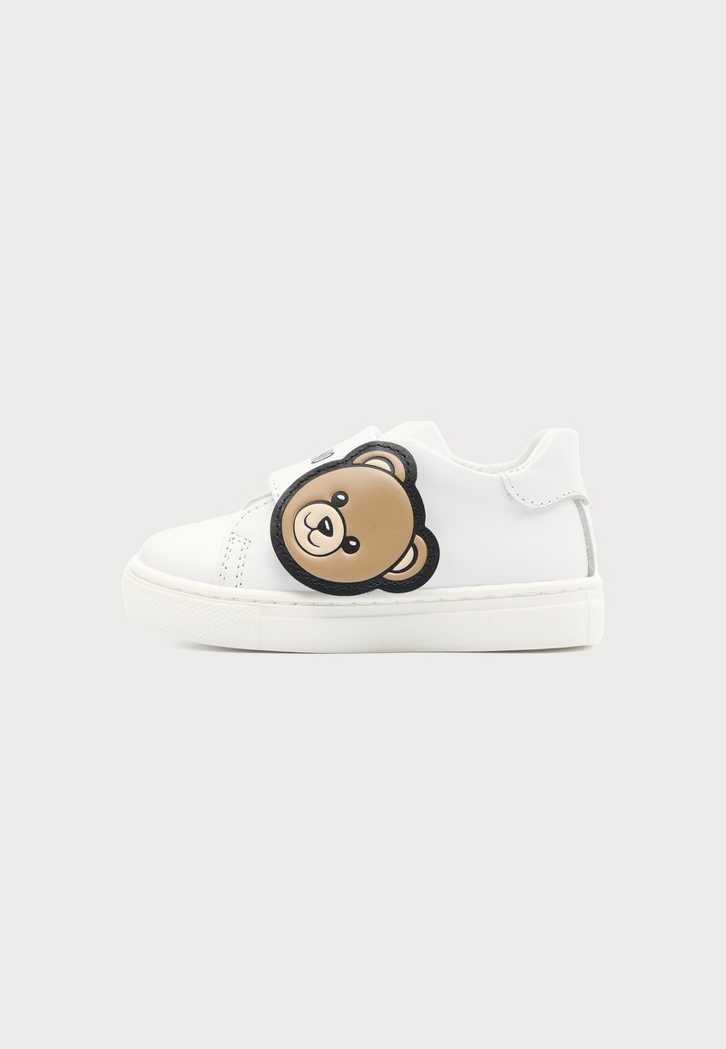 Baskets blanches pour enfants avec un patch en forme de visage d’ourson marron sur le côté et une semelle blanche épaisse sur un fond uni.