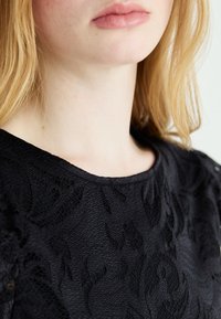 Juodas tekstūruotas viršus su gėlių raštu, plačia iškirpte ir lygiu paviršiumi, pabrėžiantis sudėtingus dizaino elementus ir sluoksniuotą audinį.