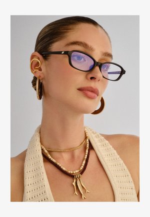 Donna con capelli lisci che indossa occhiali con montatura nera, anello auricolare d'oro, grandi orecchini a cerchio e collane a strati con ciondoli a forma di pesce.