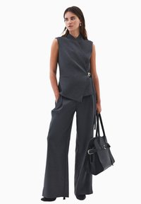 Grå ærmeløs blazer med skræddersyet pasform og asymmetrisk front, parret med brede grå bukser. Holder en sort håndtaske med spænde-detalje.