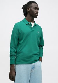 Polo vert à manches longues avec col, doté de deux boutons au niveau du cou et d'un petit logo brodé sur la poitrine, associé à un jean bleu clair.